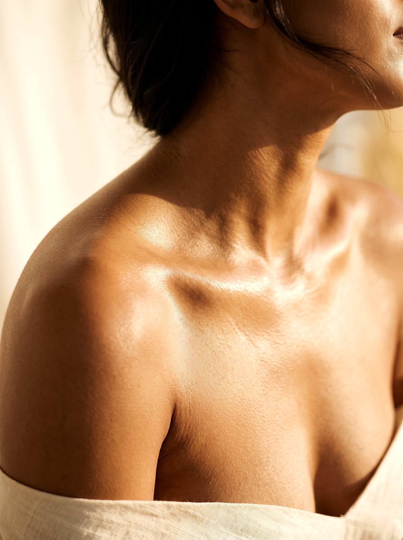 Luminous glass-skin close-up of a woman's shoulder, collarbone and neck in warm golden light