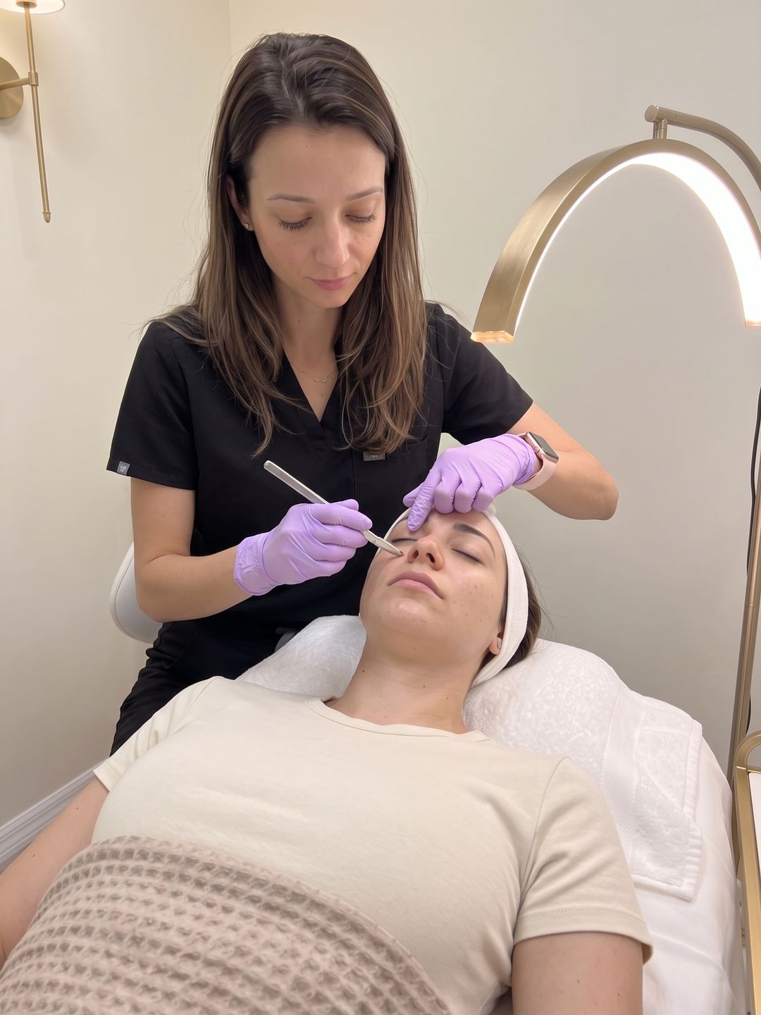 Gloved aesthetician holding a small sterile dermaplaning blade at a shallow angle against a relaxed woman's cheek on a facial bed