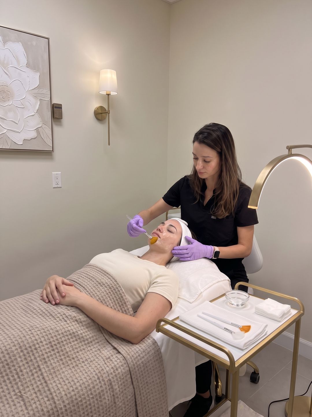 Nurse applying chemical peel solution with a fan brush to a relaxed woman's cheek on a facial bed