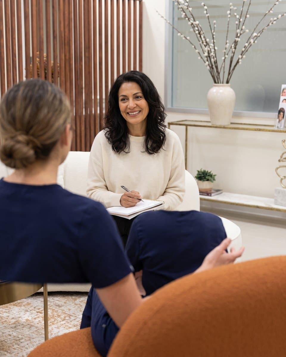A woman talking with a clinician seen from behind during a menopause consultation, notebook in hand