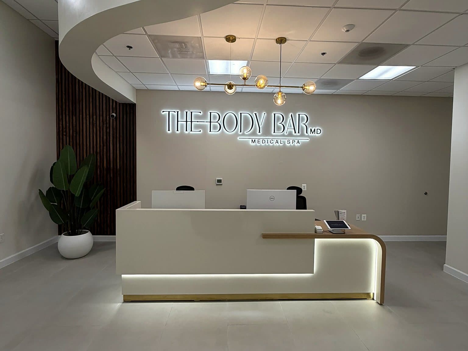 The wide front desk and reception of The Body Bar MD with its illuminated logo and sculpted ceiling